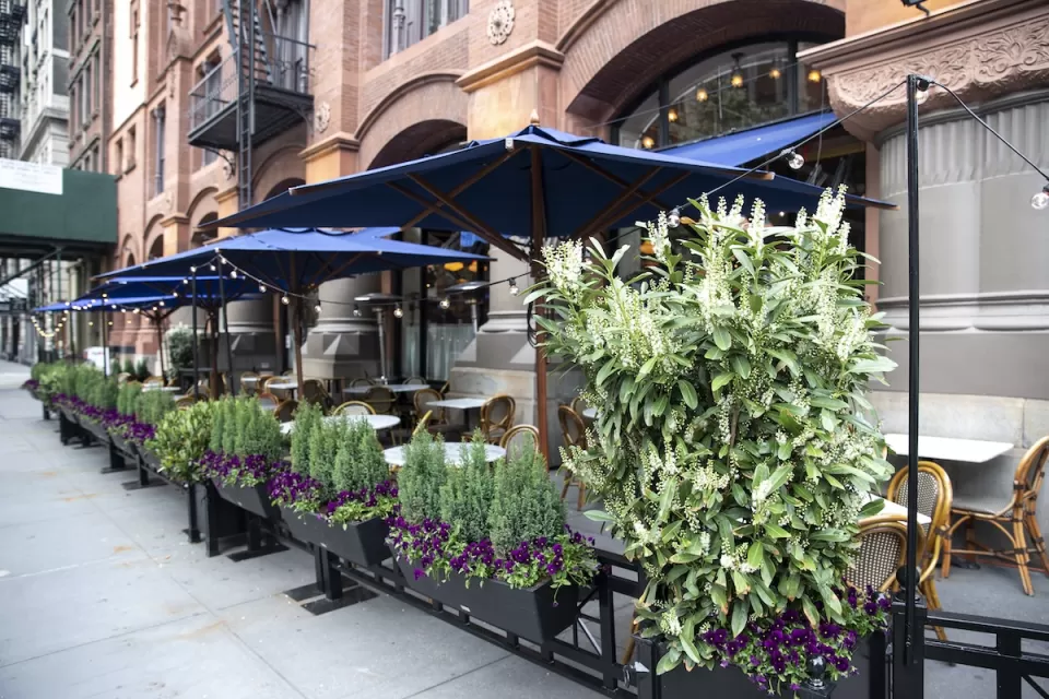 Outdoor dining patio with tables and boxes of flowers surrounding it on New York City sidewalk.